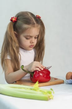 Girl playing in a cook. Girl cuts the knife red pepper. In the foreground lies celery. On the right are the bottles with the sauce, banks with green peas and corn. Studio. Light background.