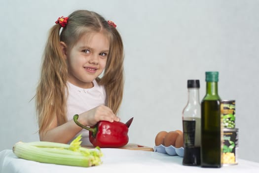 Girl playing in a cook. Girl cuts the knife red pepper. In the foreground lies celery. On the right are the bottles with the sauce, banks with green peas and corn. Studio. Light background.