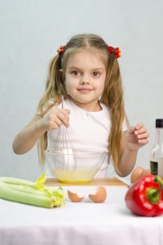Girl playing in a cook. Girl churn whisk the eggs in a glass bowl. At the forefront of the shell, celery, pepper. Studio. Light background.