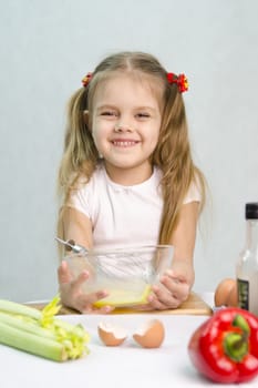 Girl playing in a cook. Girl churn whisk the eggs in a glass bowl. At the forefront of the shell, celery, pepper. Studio. Light background.