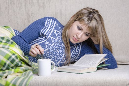 Girl sitting on the couch reading a book. Standing next to a Cup of tea, girl prevents sugar with a spoon. A girl dressed in a warm sweater and covered her warm blanket