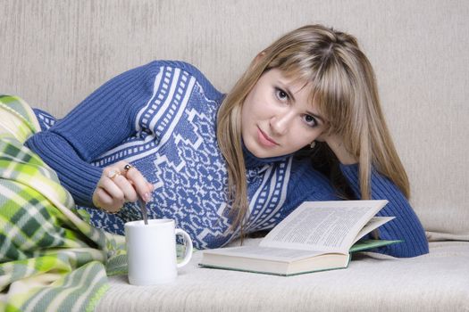 Girl sitting on the couch reading a book. Standing next to a Cup of tea, girl prevents sugar with a spoon. A girl dressed in a warm sweater and covered her warm blanket
