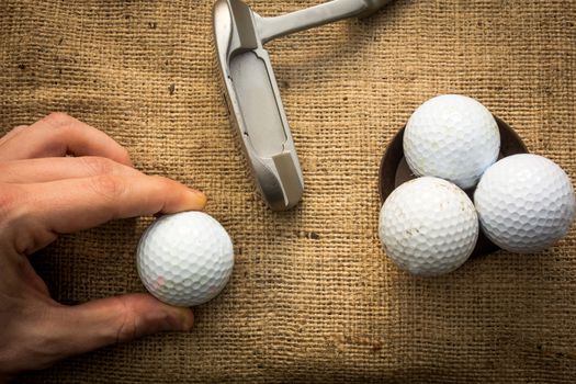 A hand holding a golf ball near a putter and three other golf balls