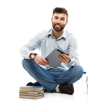 Young bearded smiling man holding a tablet with books and a bottle of water sitting on a white background