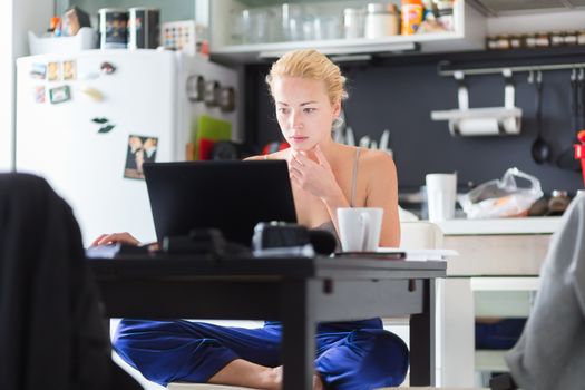 Female freelancer in her casual home clothing working remotly from her dining table in the morning. Home kitchen in the background.