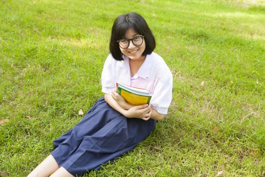 Schoolgirl holding books and smiling. Sitting on grass in the park.