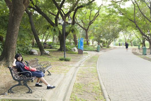 Girl wearing glasses sitting on the bench. Relax under large tree And along corridor within the park.