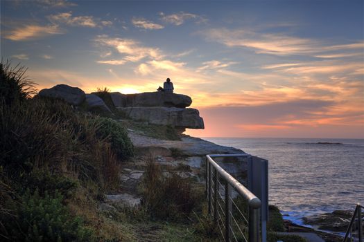 A woman and her dog watch the sunrise from a high rock at Coogee North side headland.  The best way to start the day