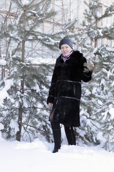 The cheerful woman in a black fur coat costs at snow-covered pines in park
