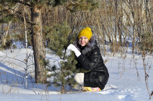 The joyful young woman sits near a pine in park in the winter