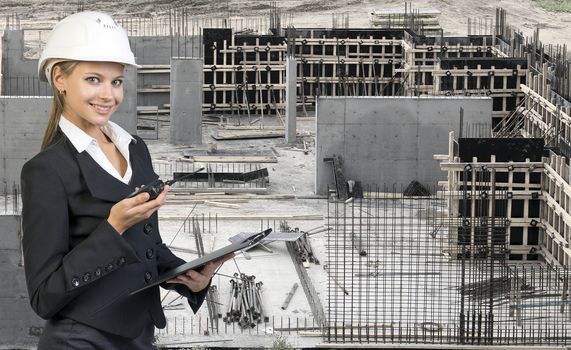 Beautiful businesswoman in suit and helmet holding paper holder and portable radio. Construction site in background. Building concept