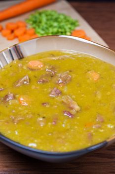 A bowl of fresh split pea soup with carrots, onions, and ham plus ingredients in the backgound.