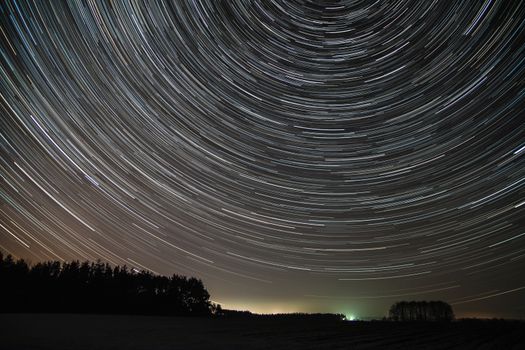 The movement of stars around the pole star over the field. Field covered with snow. City lights can be seen on the horizon.