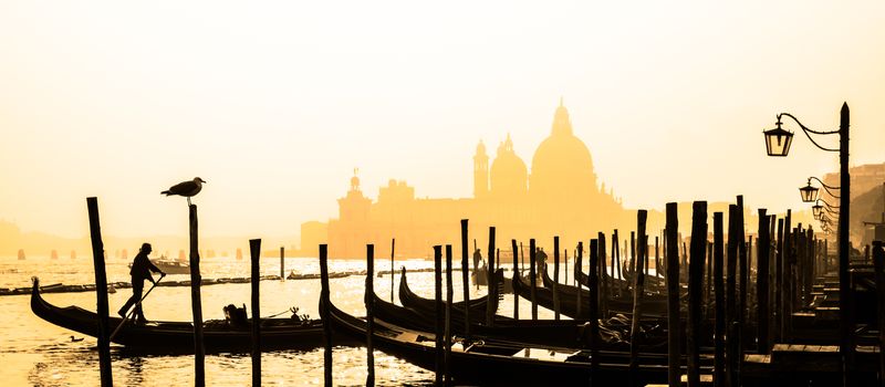 Romantic Italian city of Venice, a World Heritage Site: traditional Venetian wooden boats, gondolier and Roman Catholic church Basilica di Santa Maria della Salute in the misty background.