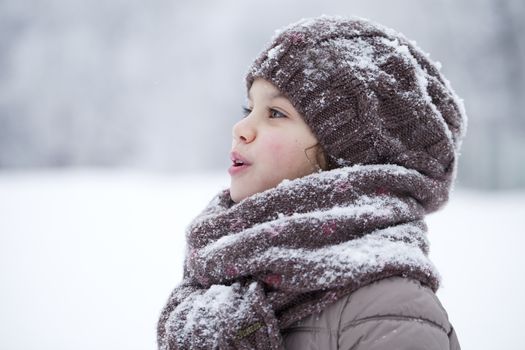 Close-up portrait of a little girl in brown jacket and knit scarf and hat on a background of a snow park