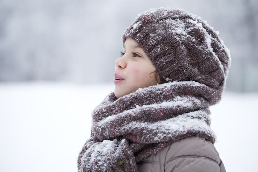 Close-up portrait of a little girl in brown jacket and knit scarf and hat on a background of a snow park