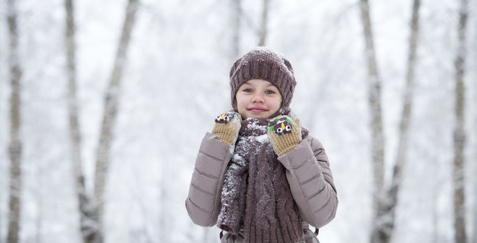 Close-up portrait of a little girl in brown jacket and knit scarf and hat on a background of a snow park