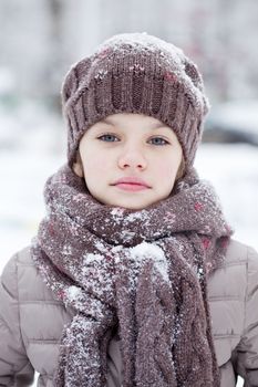 Close-up portrait of a little girl in brown jacket and knit scarf and hat on a background of a snow park