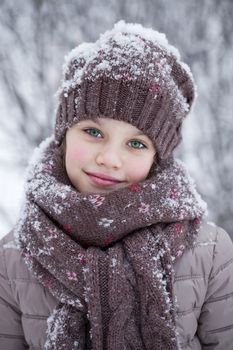 Close-up portrait of a little girl in brown jacket and knit scarf and hat on a background of a snow park