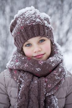 Close-up portrait of a little girl in brown jacket and knit scarf and hat on a background of a snow park