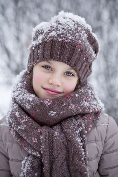 Close-up portrait of a little girl in brown jacket and knit scarf and hat on a background of a snow park