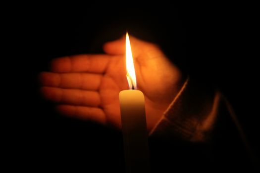 hand protects the flame from a candle burning on a dark background