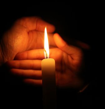 hand protects the flame from a candle burning on a dark background