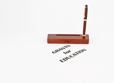 Pen in rosewood stand indicates writing and text directs to grants and proposals that can fund schools and educational costs, supplies, and projects. Copy space available around direct, still life metaphors of horizontal image.