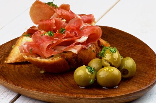 Delicious Tapas with Smoked Jamon on Garlic Bread and Heap of Green Olives on Wooden Plate closeup on White Wooden background