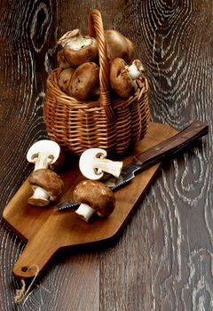 Arrangement of Gourmet Raw Portabello Mushrooms in Wicker Basket with Table Knife on Cutting Board closeup on Dark Wooden background