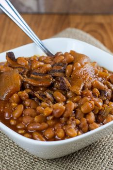 Rustic baked beans in a white bowl on a burlap placemat and wooden table.