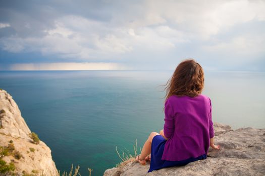 Young beautiful girl sitting on a rock and looking at sea.