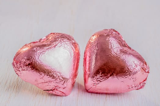 Two chocolate hearts, in pink wrapping foil, close together on a white table.