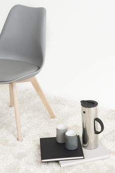Serving of coffee alongside a modern molded chair with two empty mugs and an elegant tall flask resting on two hardcover books on a white carpet, with copyspace
