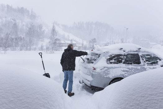 A man is shoveling snow of a car. It`s snowing and snowflakes are visible