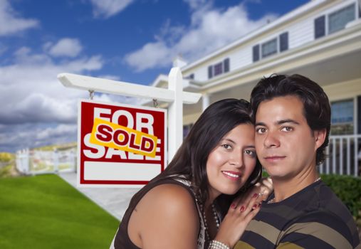 Young Happy Hispanic Couple in Front of Their New Home and Sold For Sale Real Estate Sign.