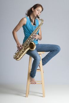 Pretty brunette musician playing a tenor saxophone while seated on a wooden stool