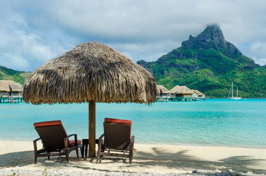 Two sunlounger chairs under a thatched parasol on a white sand beach with a view on the lagoon and the tropical island of Bora Bora, near Tahiti, in French Polynesia.