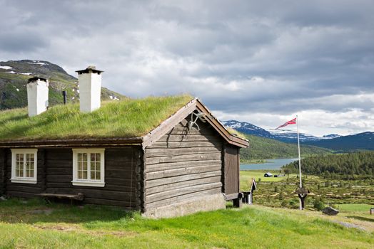 A sod roof log cabin with grass on top and two chimneys with a view over a lake in a valley in Jotunheimen national park, Norway.
