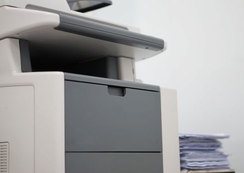 White copiers located in the office, side of the machine has a pile of papers.                             