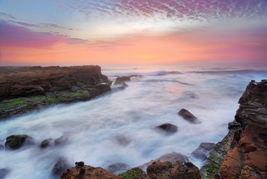 Break of dawn the rosy flamingo feathered sunrise skies compete for attention with turbulent rushing ocean flows over tidal rocks