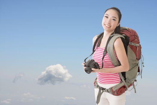 Happy smiling Asian young female backpacker with camera standing in front of blue sky.