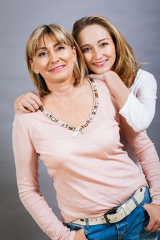 Playful beautiful young mother and her teenage daughter posing together with the young girl peeking out to the side with a happy grin, isolated on a grey studio background