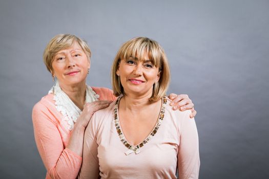 Attractive stylish blond senior lady with her beautiful middle-aged daughter posing together with her hands on her shoulders smiling at the camera on a grey studio background