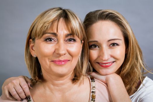 Playful beautiful young mother and her teenage daughter posing together with the young girl peeking out to the side with a happy grin, isolated on a grey studio background