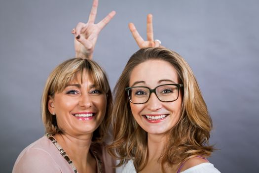 Playful beautiful young mother and her teenage daughter posing together with the young girl peeking out to the side with a happy grin, isolated on a grey studio background