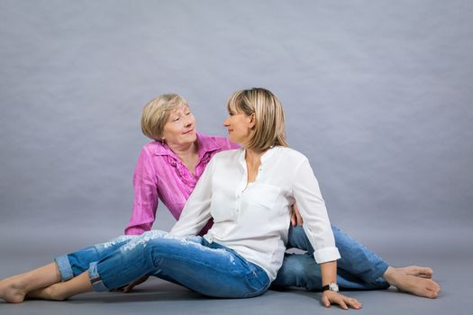 Attractive stylish blond senior lady with her beautiful middle-aged daughter posing together with her hands on her shoulders smiling at the camera on a grey studio background