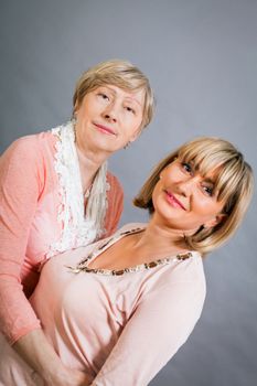 Attractive stylish blond senior lady with her beautiful middle-aged daughter posing together with her hands on her shoulders smiling at the camera on a grey studio background