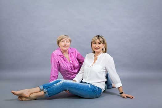 Attractive stylish blond senior lady with her beautiful middle-aged daughter posing together with her hands on her shoulders smiling at the camera on a grey studio background