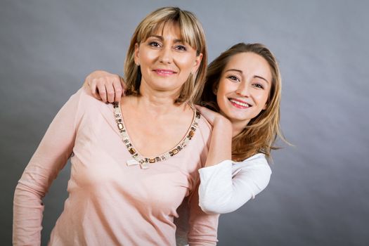 Playful beautiful young mother and her teenage daughter posing together with the young girl peeking out to the side with a happy grin, isolated on a grey studio background